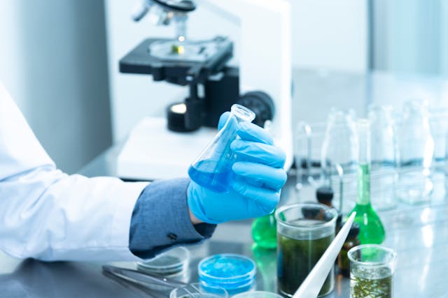 Scientist in gloves holding a flask with blue liquid on a lab bench with glassware and a microscope.