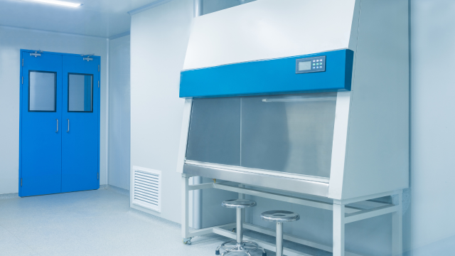 A clean, modern laboratory room featuring a biosafety cabinet with stools in front and bright blue double doors in the background.