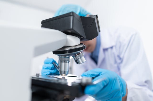 A scientist wearing blue gloves and a lab coat looks through a microscope in a laboratory setting.