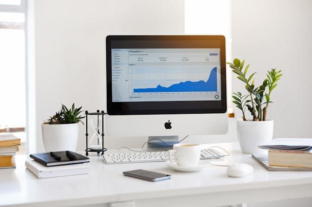 A modern workspace featuring a white desk with an Apple iMac displaying a graph with rising data. The desk is neatly organized with a cup of coffee, a smartphone, books, an hourglass, and small potted plants. Natural light streams in, creating a bright and minimalistic office setting.