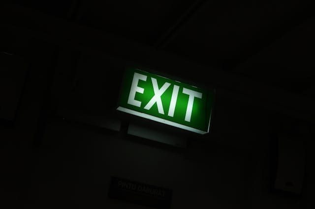 A glowing green "EXIT" sign is illuminated in a dimly lit indoor space, indicating an emergency exit. The background is dark, making the sign stand out prominently.